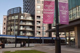 The ION Light Rail drive by high rise buildings in Downtonw Kitchener, In the froreground are banners reading "welcome to Downtown kitchener" and "have an awesome day.
