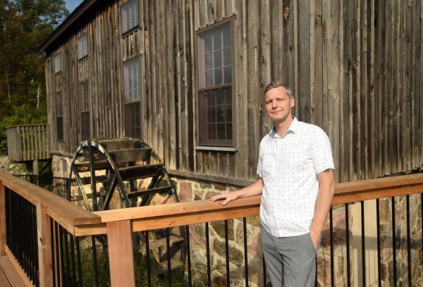 A man stands in front of a historical wooden building and water wheel on a sunny day